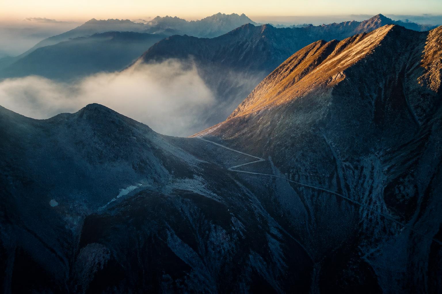 A faint trail crossing a high mountain saddle at dawn, ridgelines receding into cloud